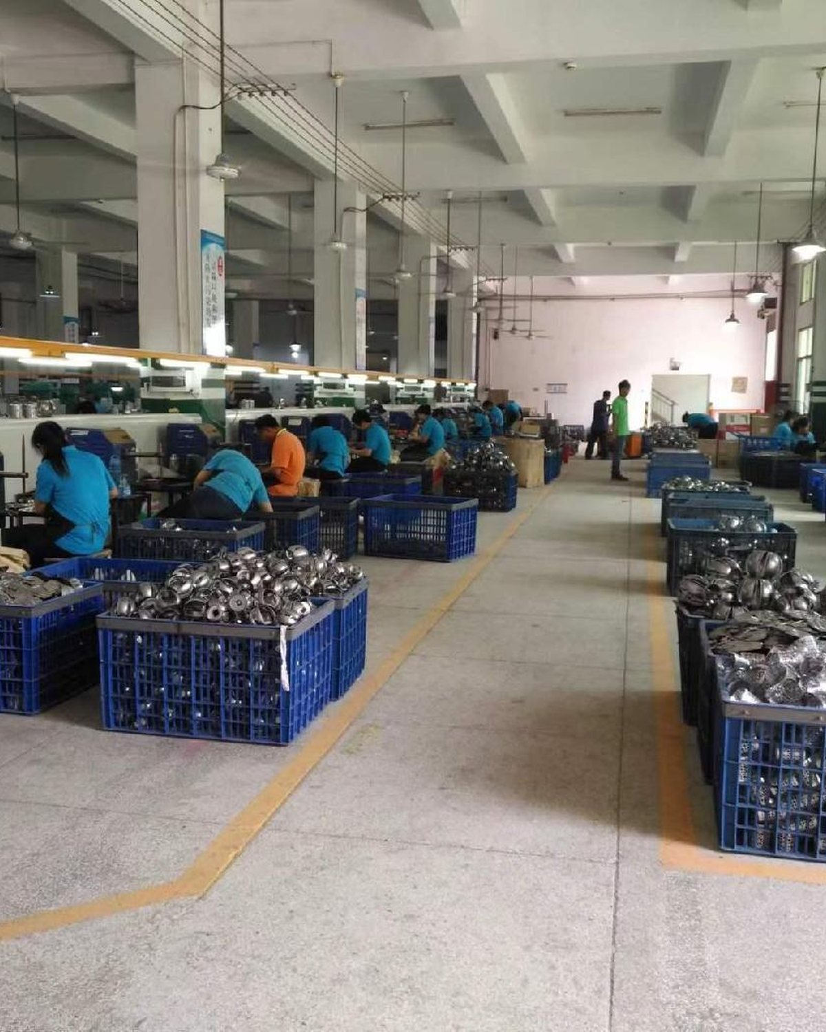 A pressing and forming hall: rows of workers at presses with blue crates of stamped raw metal parts stacked along the aisle